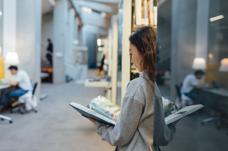 Person stands beside a bookshelf reading an open book in a modern library, with others studying at desks in the background.