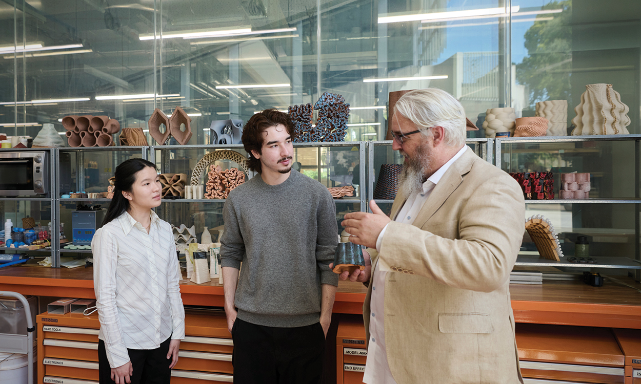 An instructor in a tan jacket gesturing and speaking with two young adults in a workshop studio.