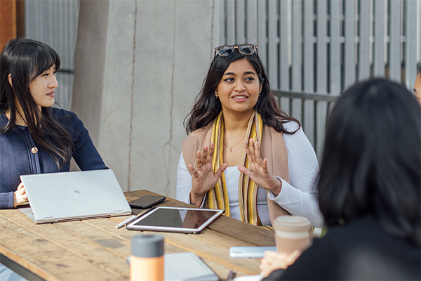 a group of four students sitting outside at a table with laptops and tablets in conversation