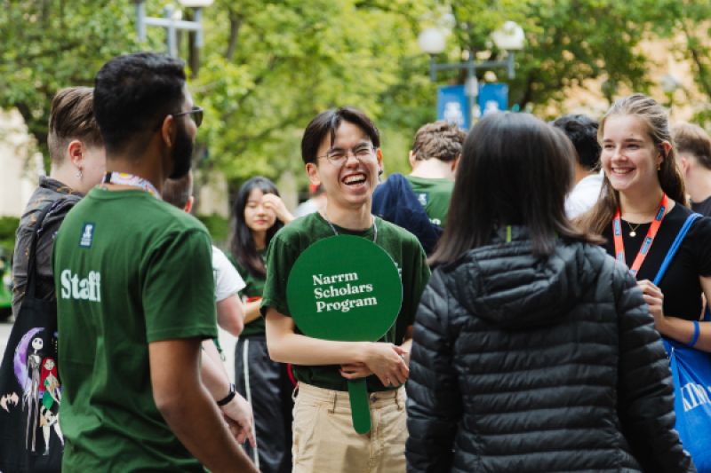 Group of young people talking outdoors, with a round green sign for the Naarm Scholars Program