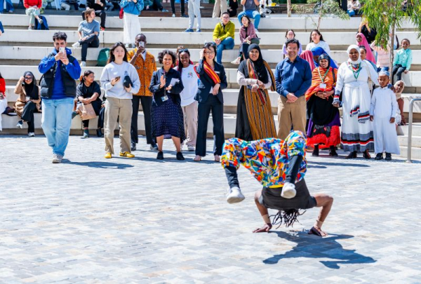 A performer in colourful African clothing balances in a handstand on an open courtyard, with a crowd gathered behind them.