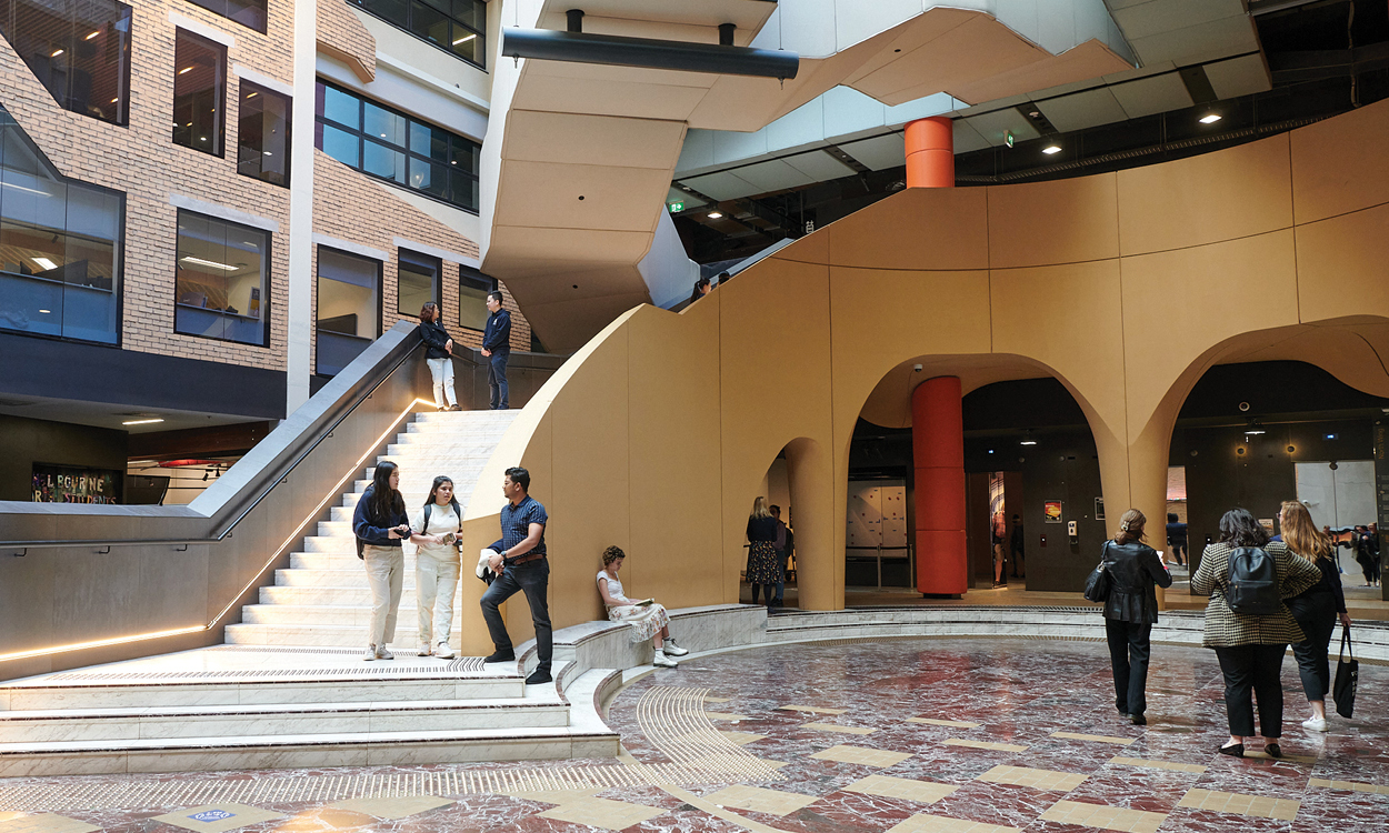 Students chatting on a staircase in a University of Melbourne building atrium, with archways and a circular marble floor in the background.