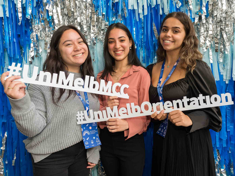 Three students standing in front of a sparkly photo wall, holding props which say #UniMelbMCC and #UniMelbOrientation