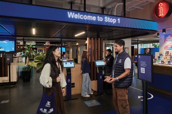 A staff member speaks with a visitor beneath a sign that reads “Welcome to Stop 1,” surrounded by digital kiosks and people moving through the space.