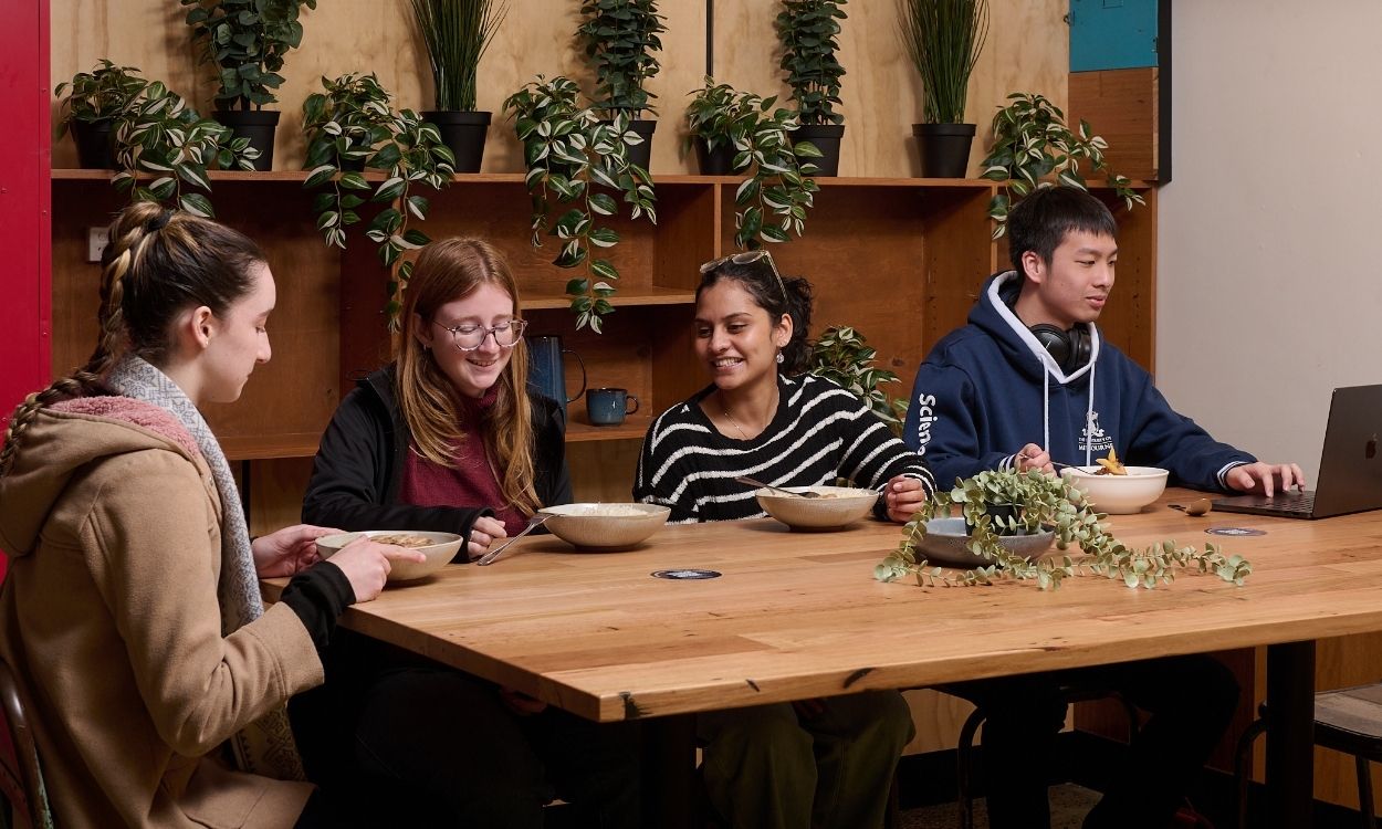 Students sitting together at a wooden table in a casual indoor campus setting, eating from bowls and chatting, with one student using a laptop. Green plants line shelves behind them, creating a relaxed, social atmosphere.