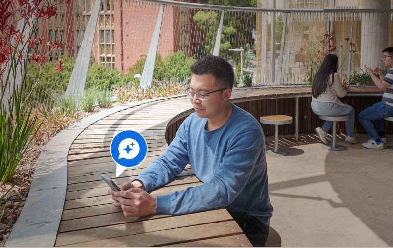 A person in a blue sweater sits on an outdoor wooden bench with a smartphone showing a blue AI sparkle icon.