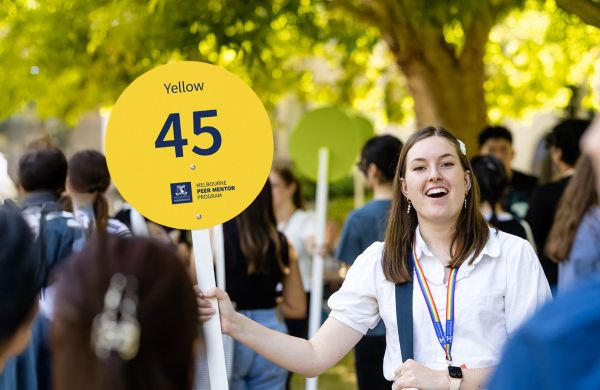 A smiling woman in a crowd holds a yellow sign for the Melbourne Peer Mentor Program.