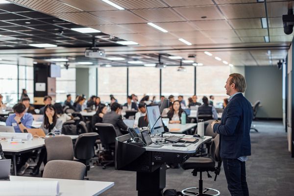 A lecturer speaks to a large room of students seated at collaborative tables with laptops and documents.