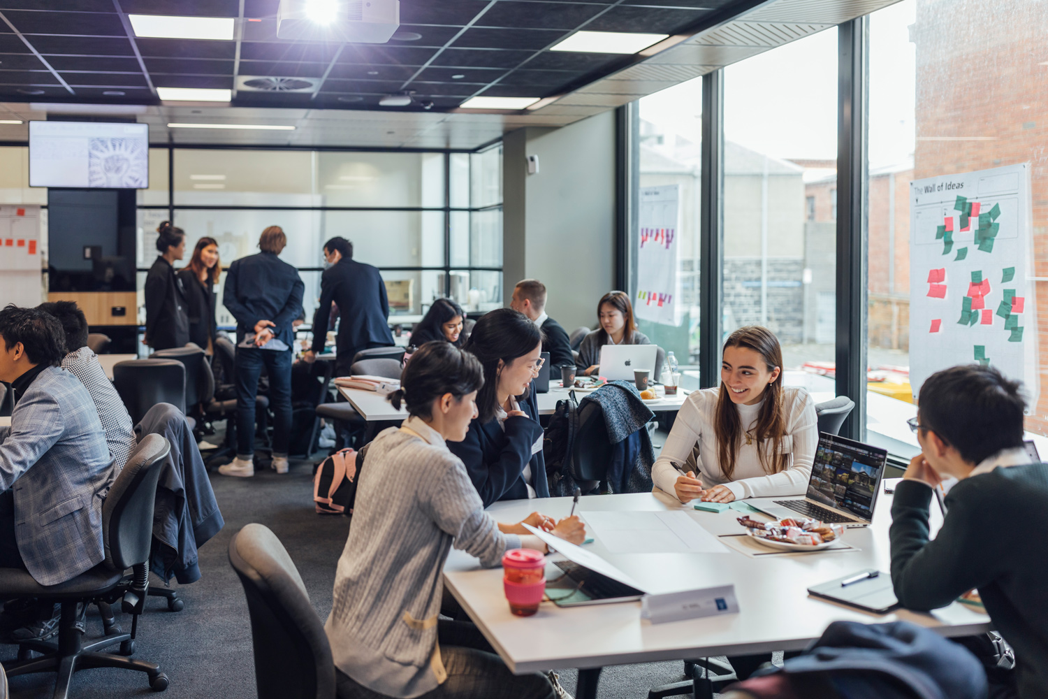 Uni students collaborate around a table in a sunlit lounge with laptops and notebooks.