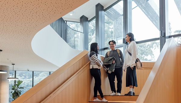 Three students stand on a staircase landing and talk together.