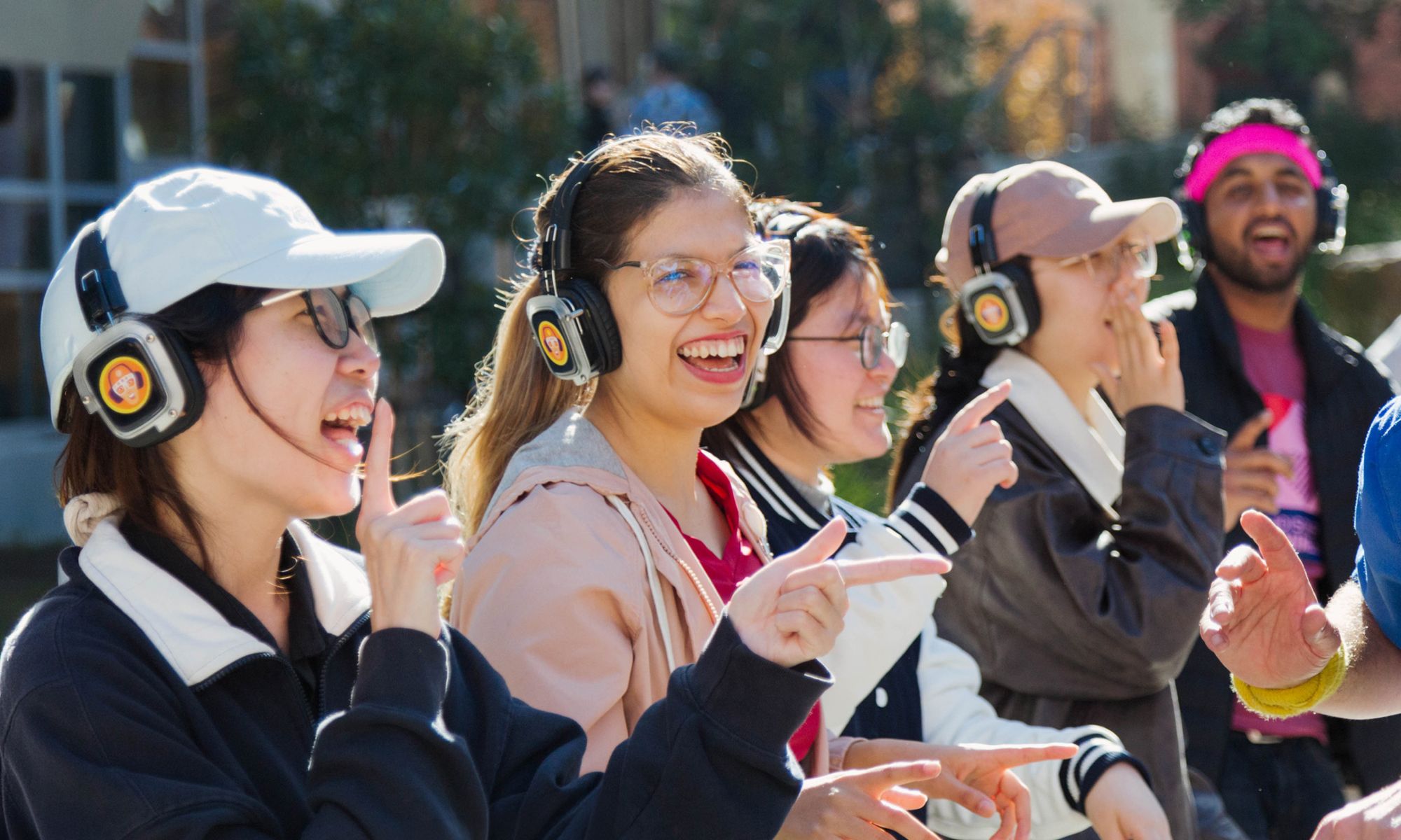 A group of people wearing large over‑ear headphones stand outdoors in daylight, all pointing in various directions as part of an activity. Trees and buildings are visible softly in the background.
