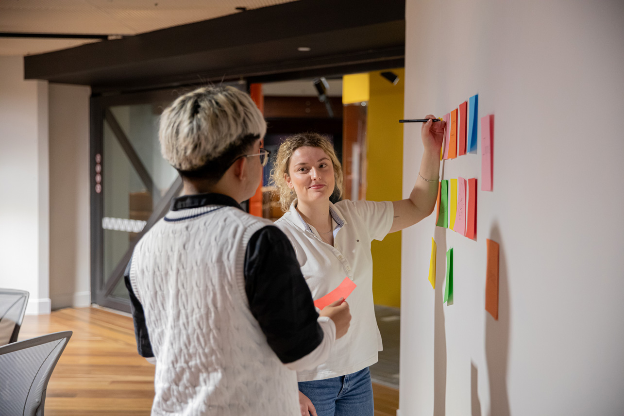 Two people collaborate, one writing on a wall with colorful notes.