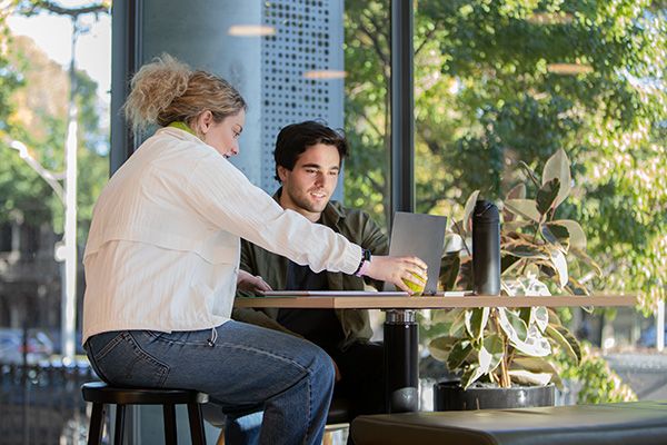 Two students collaborate at a table by a large window with a view of green trees.
