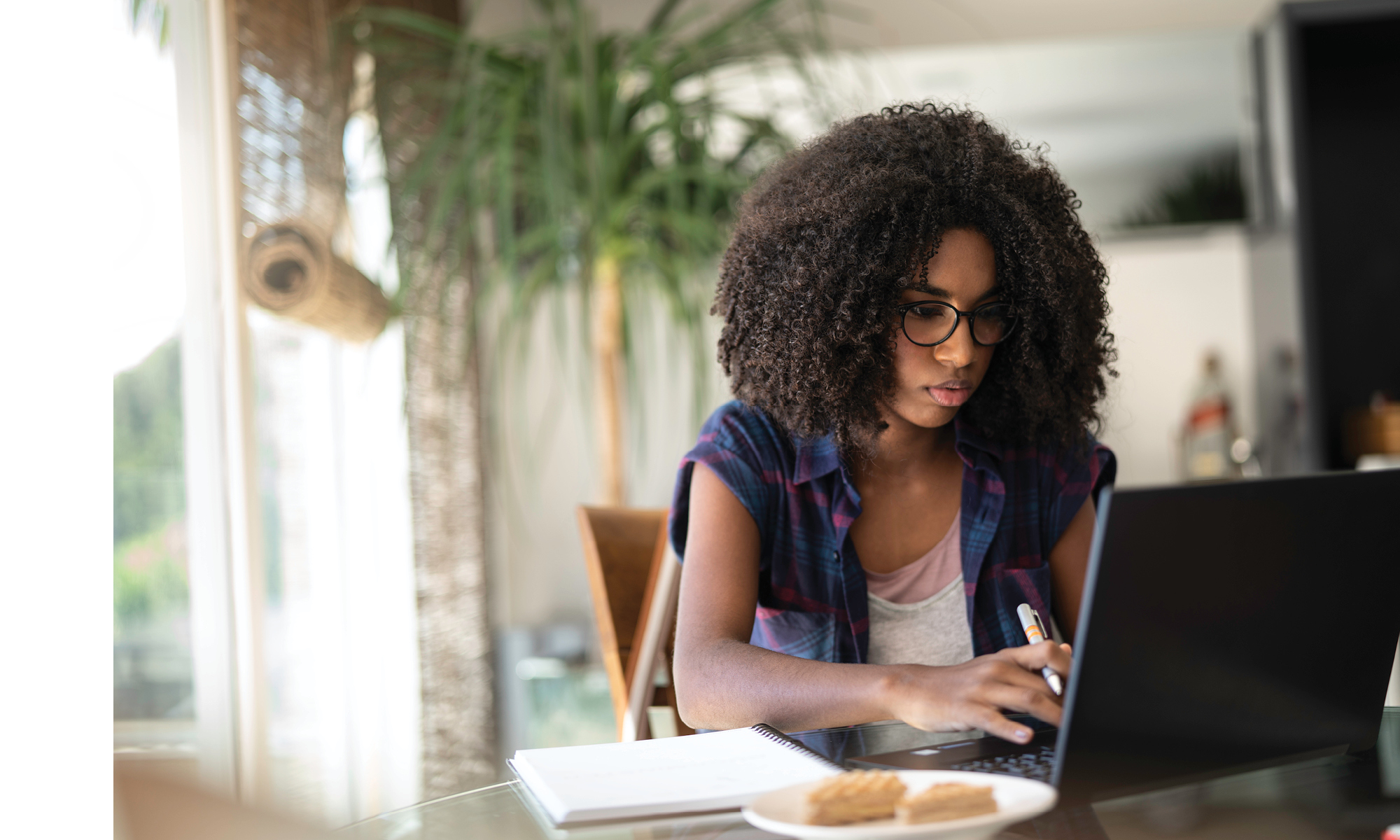Person working on a laptop at a dining table with a notebook and pen nearby.