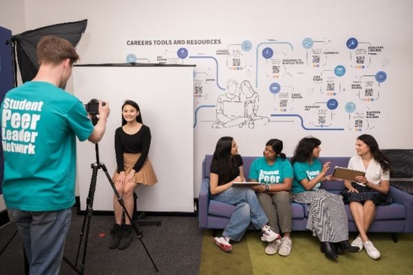 A Student Peer Leader Network member photographs a student in front of a white backdrop while other students chat on a nearby couch.