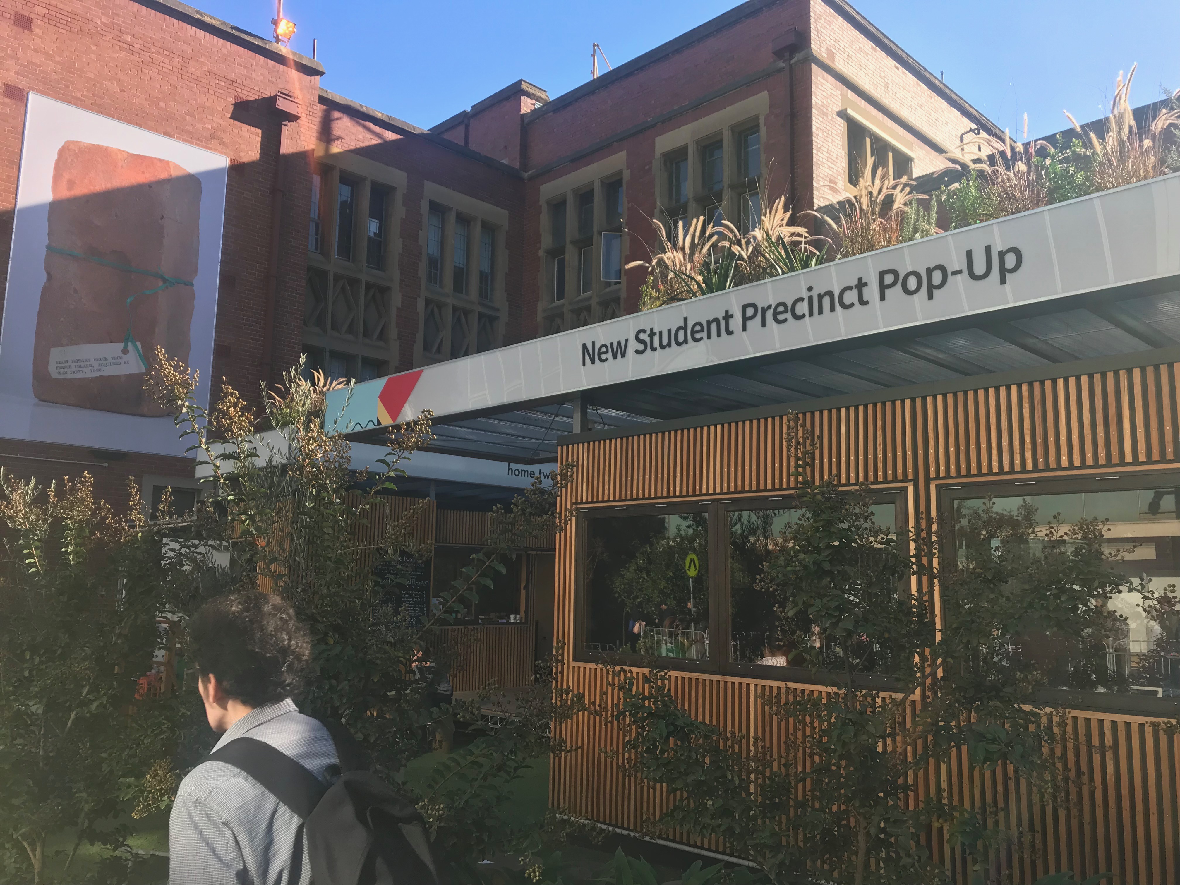 An outdoor study pod at the Mon-Pop site. The pod is a large, rectangular struture, covered in green grasses and made of hard, deep brown wood. A white sign above the structure reads "New Student Precinct Pop-Up. A student in a grey hoodie is walking by the structure