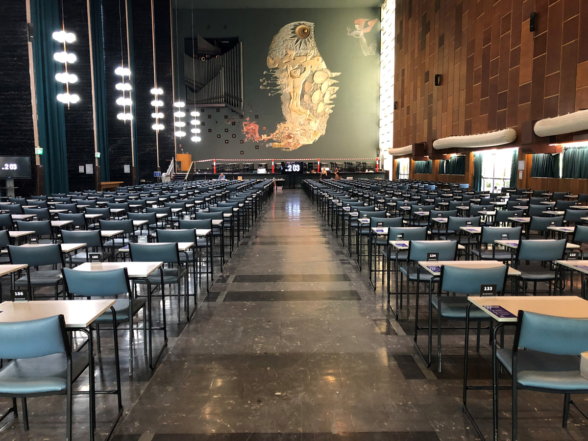 Many rows of individual chairs and tables for exams, facing a huge mural in Wilson Hall.
