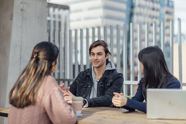 Three students engage in a conversation while sitting at a wooden table on an outdoor terrace.