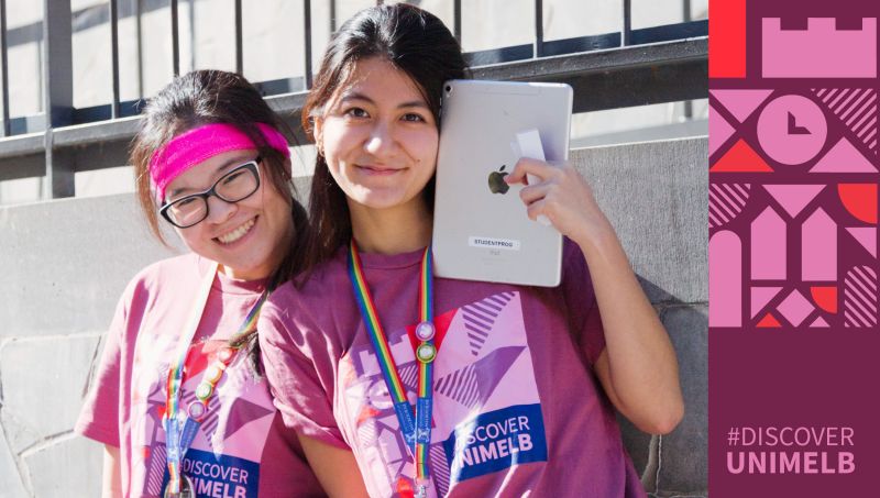 Two people wearing matching pink “Discover Unimelb” T‑shirts and lanyards pose outdoors; one holds up a tablet. A pink and purple graphic panel on the right features geometric shapes and the text “#DISCOVER UNIMELB.”