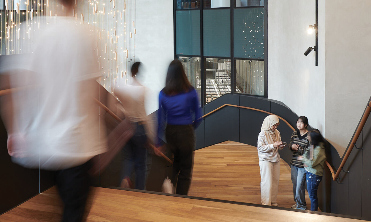 Motion-blurred people walk up a curved wooden staircase in a modern building while three people stand together on the landing near large windows.