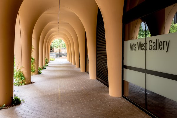A long arched walkway with smooth, sand‑coloured columns leads toward a green outdoor area. A glass wall on the right displays the text “Arts West Gallery.”