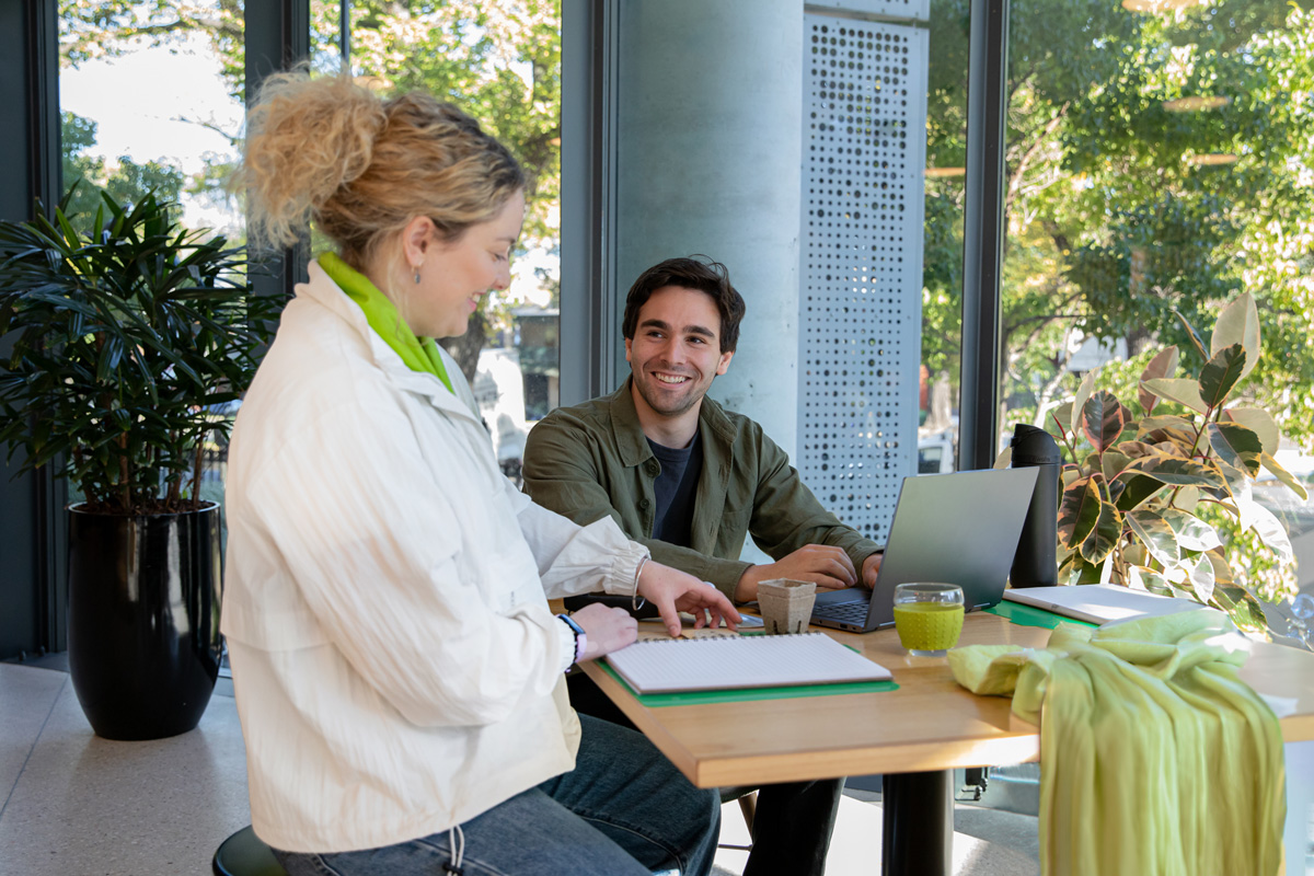 Two people talk at a table with a laptop and plants.