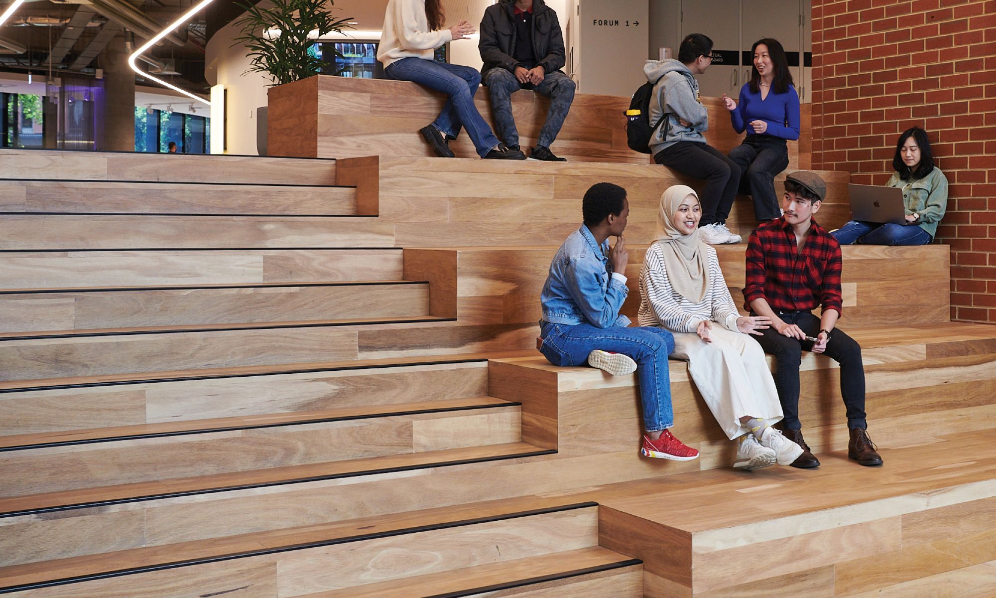 A group of students are talking with peers while sitting in an open plan lecture theatre with wooden steps.