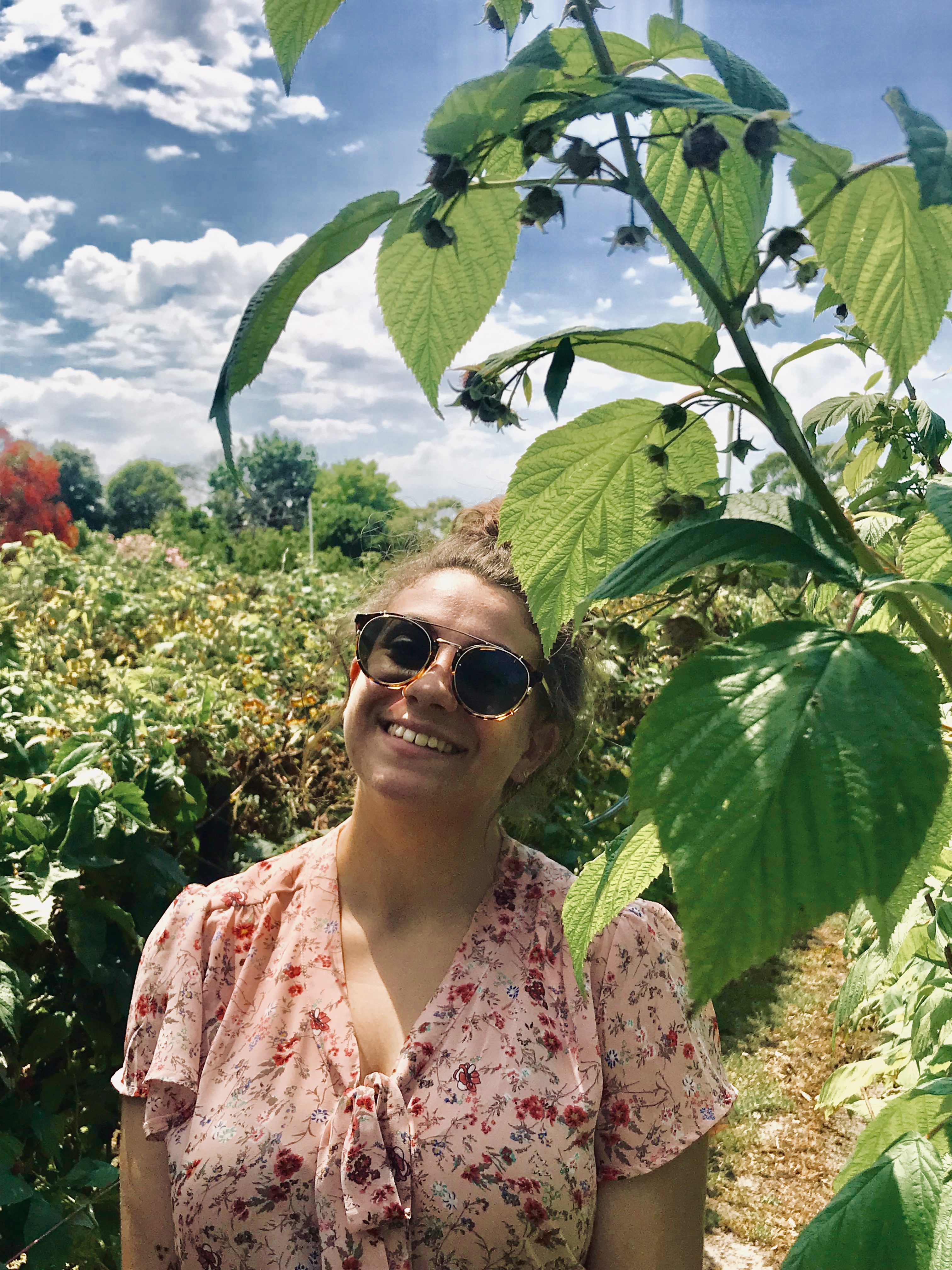 Yasmin is standing in a field of sunflowers, smiling at the sky in a pink shirt