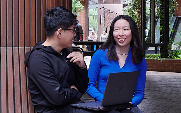 Two people sit together outdoors on a wooden bench, talking while one of them uses a laptop. Trees and brick walkways surround the area.