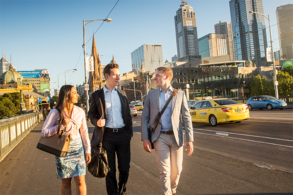 three professional looking students walking outside along bridge in a conversation with the sun shining on them