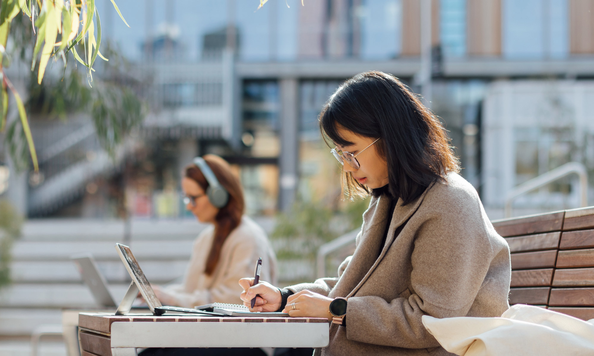 Student in a tan jacket, sitting on a bench on campus checking their results on their laptop and taking notes on a notepad.