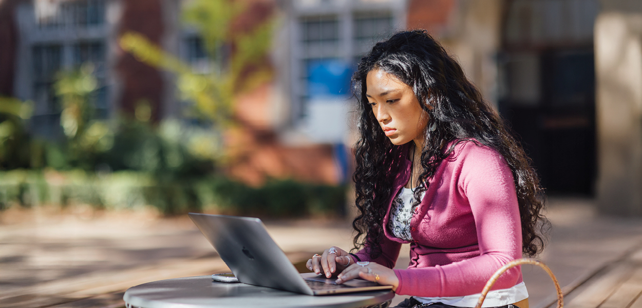 A young woman with long dark hair focuses on her laptop outdoors.