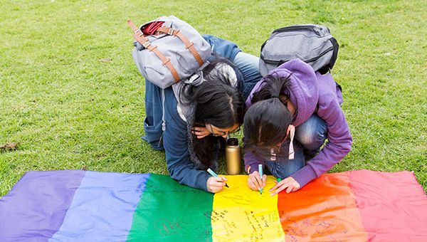 Two students are crouching down to write messages on a vinyl pride flag lying on the ground.