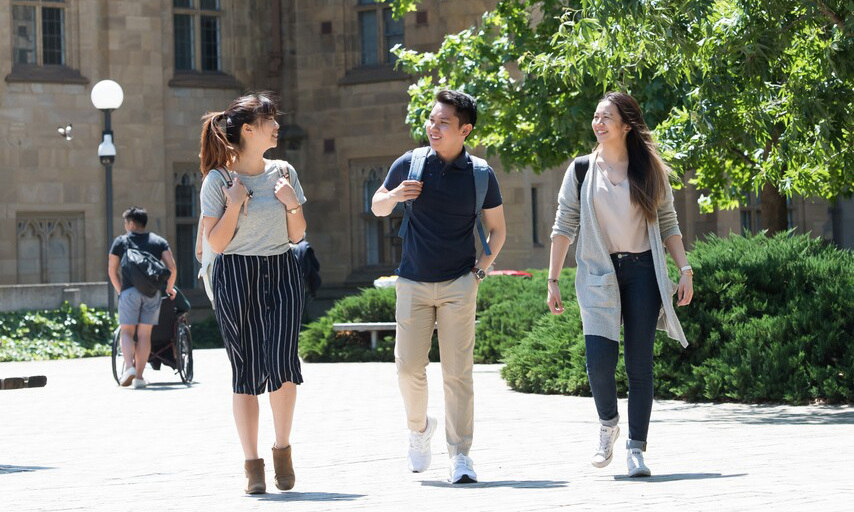 Three students chat on a sunny campus pathway; one wheelchair user is in the background.