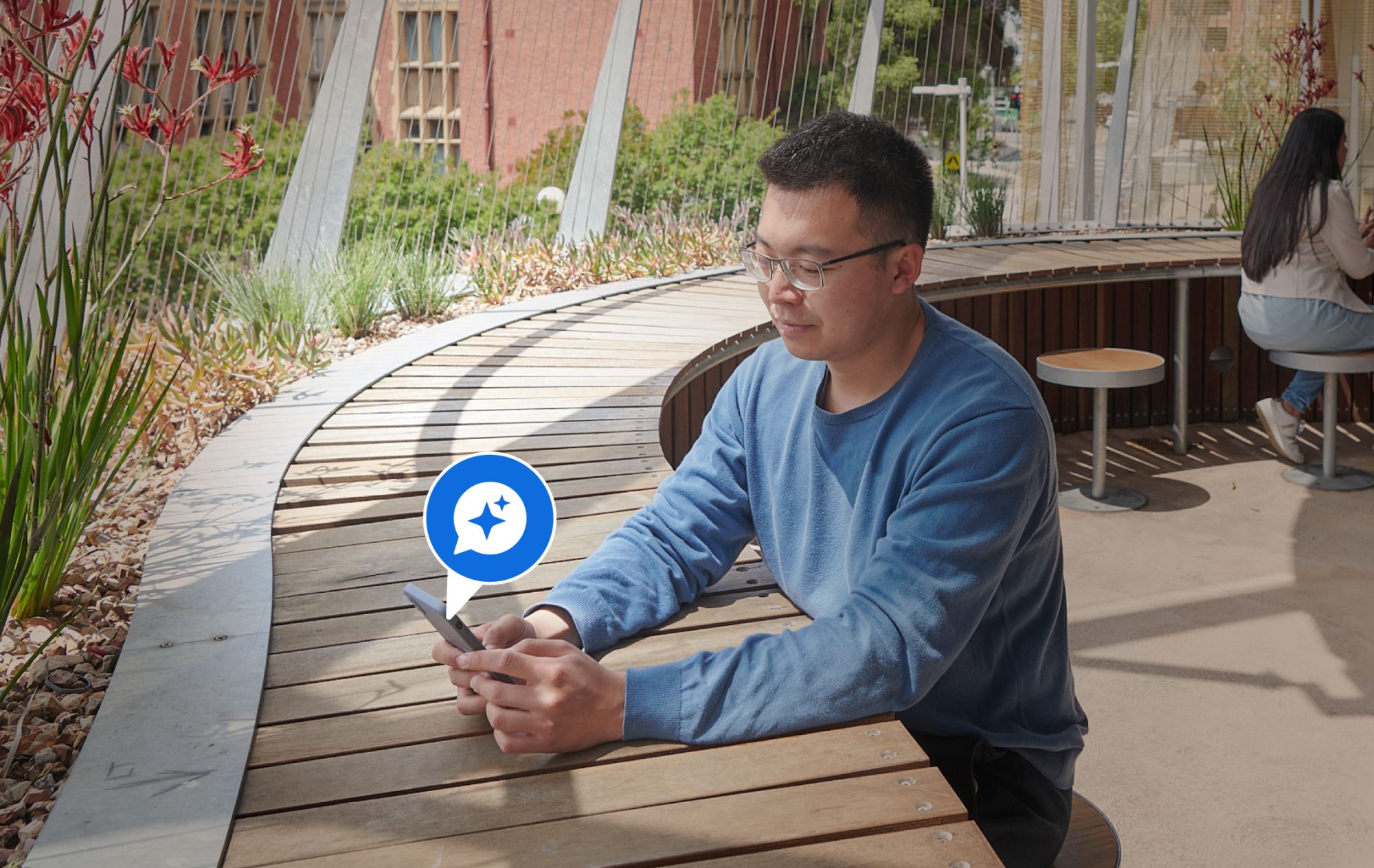 A person in a blue sweater sits on an outdoor wooden bench with a smartphone showing a blue AI sparkle icon.