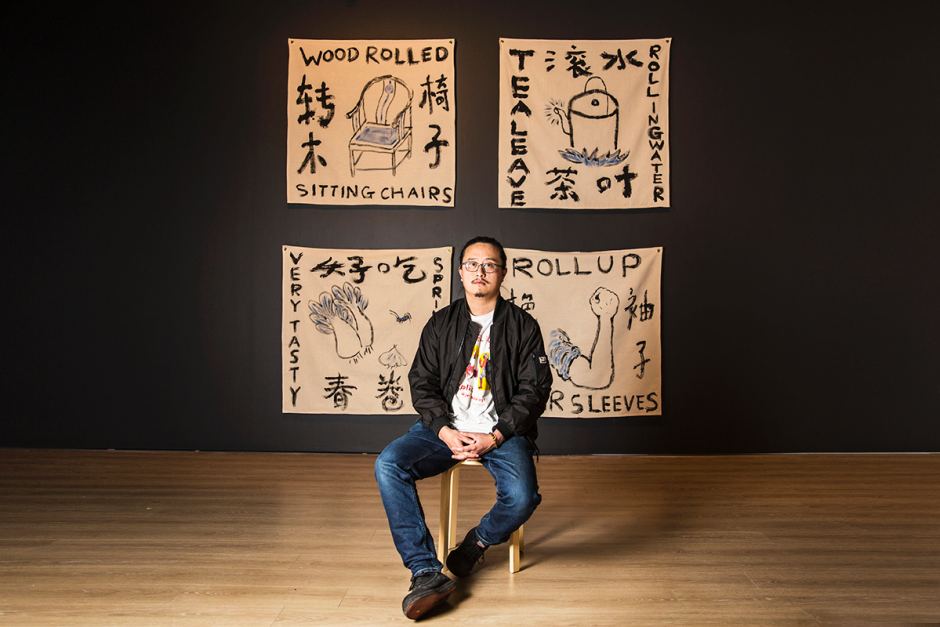 Artist Jason Phu sitting on a wooden stool infron of their artworks. Jason wears blue jeans and a black jacket. His artworks are black line drawings of everyday household objects, captioned in both English and Chinese Mandarin. There is a chair drawn with the caption 'wood rolled sitting chair', a kettle with the caption 'tea leave / rolling water', a bunch of bok choy with the caption 'very tasty', and an arm flexing it's muscle with the caption 'roll up your sleeves'